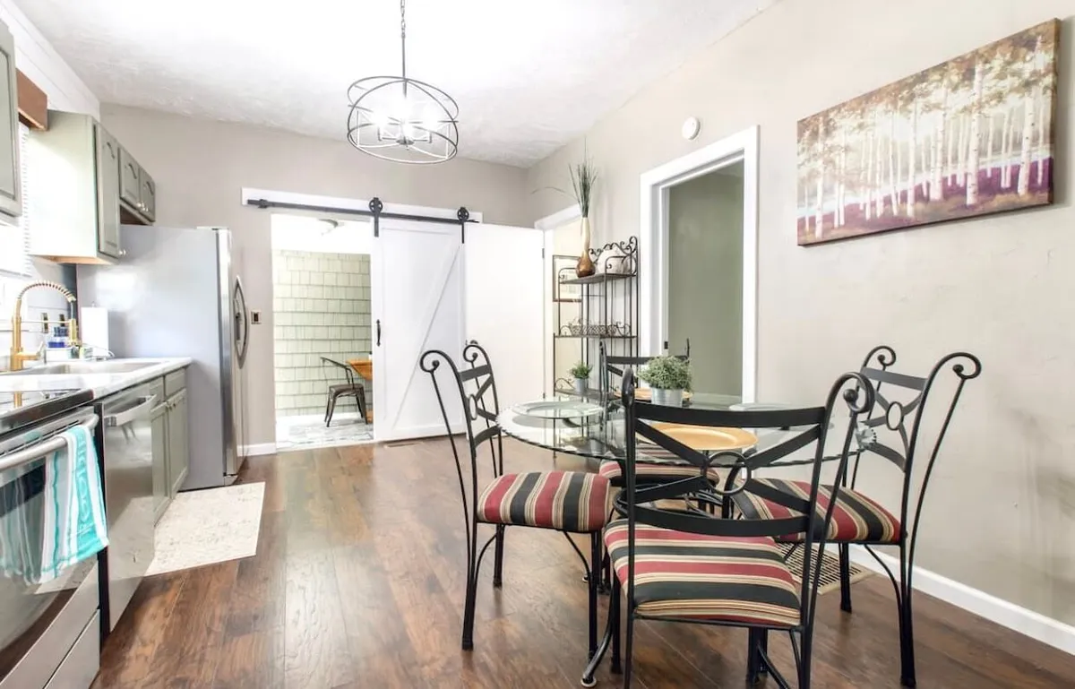 The image features a glass dining table surrounded by striped chairs, modern kitchen appliances, a barn-style sliding door, and artwork on the wall. The scene evokes a cozy, stylish, and functional space where family and friends can gather, eat, and socialize.