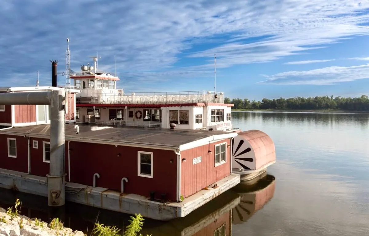 This image features a tranquil houseboat structure on the river with expansive sky above. The river’s calm surface and the relaxed scene evoke the peacefulness of riverside living and the allure of being close to water and nature.