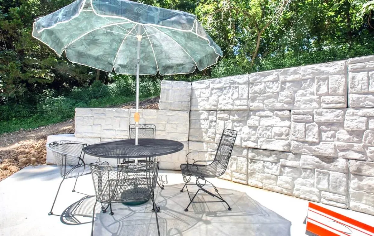 An inviting stone patio with a metal table, black chairs, and a green umbrella, set against a backdrop of greenery. The table is ideal for alfresco meals or casual gatherings, while the overall setup suggests relaxation and enjoyment of the outdoors.