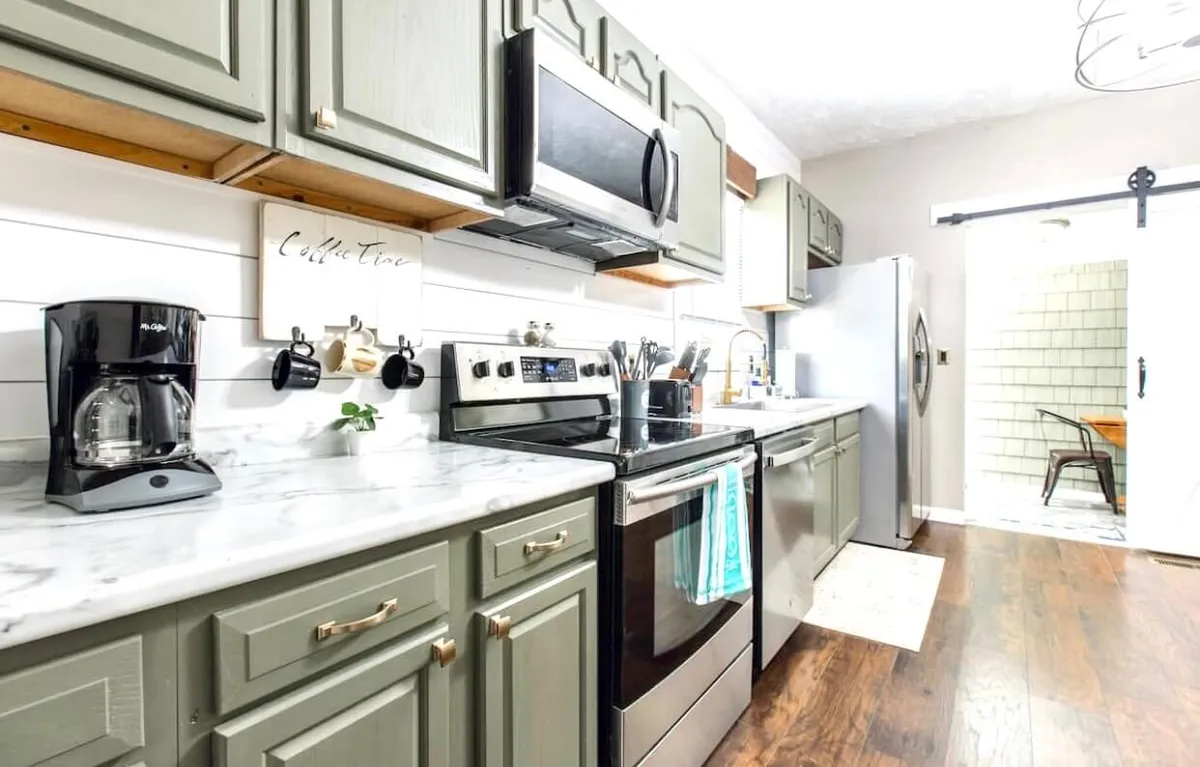 A close-up view of a kitchen with modern appliances, a marble countertop, green cabinets, a coffee maker, and open shelving. The arrangement is uncluttered and inviting, reflecting thoughtful design and practical features for meal prep and energizing mornings.