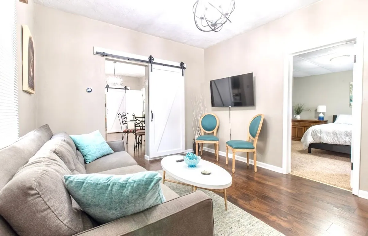 This welcoming living room scene showcases a cozy sofa, a coffee table, a TV, and an adjacent dining area. The space is arranged for socializing, relaxing, or enjoying a movie night, making it ideal for families and groups to unwind and connect. The arrangement emphasizes both comfort and versatility, echoing the 'Festival Gathering Lounge' theme.