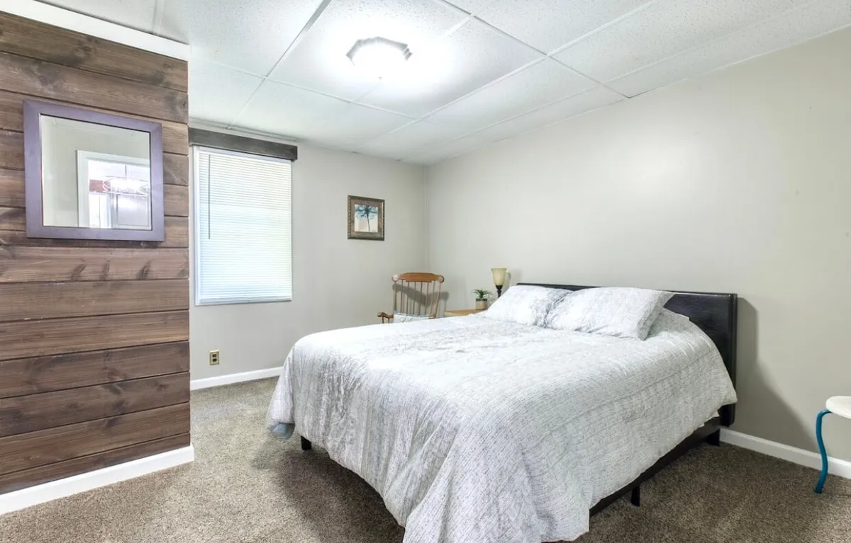 A tranquil bedroom featuring a bed with crisp white bedding, a warm wood accent wall, a window with blinds letting in natural light, and a cozy rocking chair in the corner. The scene exudes serenity and comfort, ideal for unwinding after a busy day.