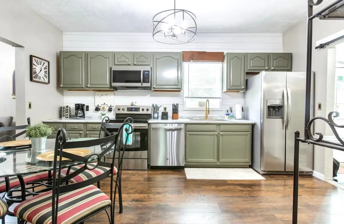 This image shows a warm, inviting kitchen with a central island, stainless steel appliances, green cabinetry, and a dining table nearby. The layout emphasizes gathering spaces for family meals and socializing, with light streaming in to enhance the welcoming mood.