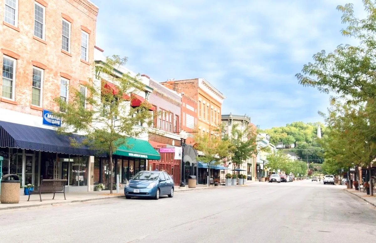 A vibrant street scene featuring historic brick buildings with colorful awnings, tree-lined sidewalks, and parked cars. The image captures the charm and energy of Hannibal’s downtown, hinting at boutique shops, outdoor dining, and areas that host lively festivals and community gatherings.
