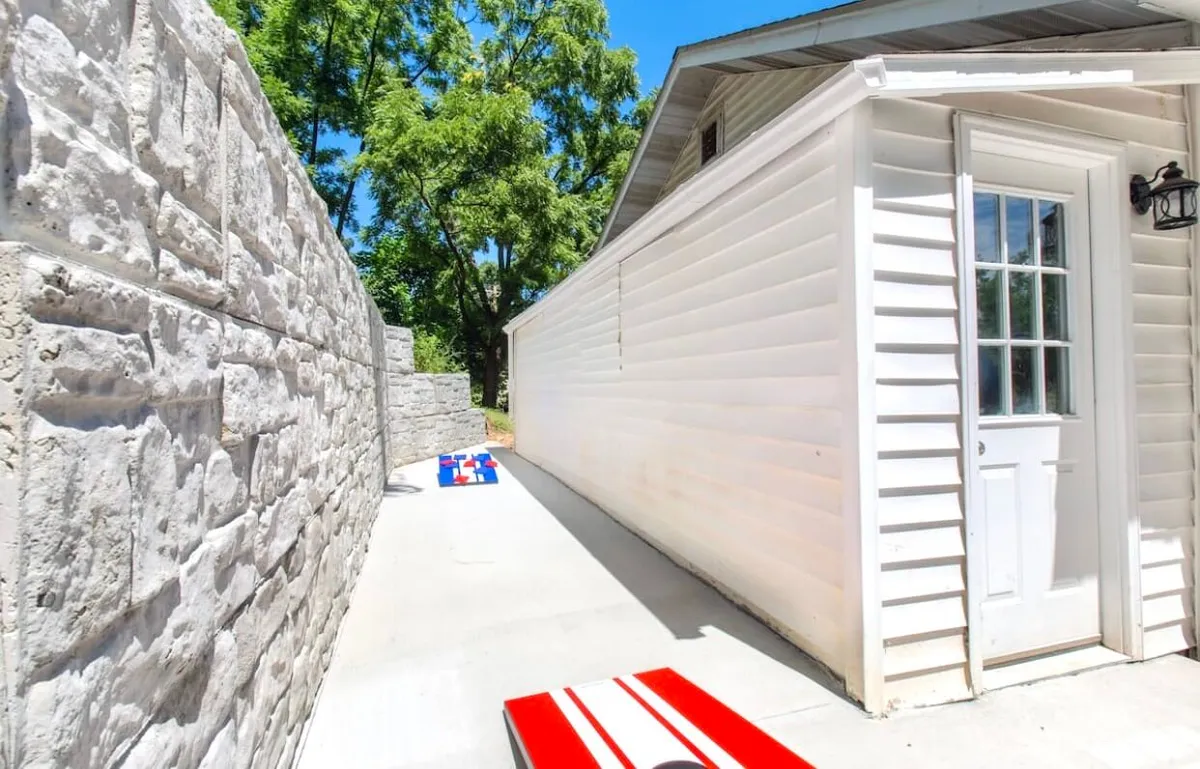 A vibrant outdoor scene featuring brightly colored cornhole boards set up on a stone patio next to a white wooden door and stone wall. Friends are mid-game, suggesting an atmosphere of casual, playful competition. The area is bathed in sunlight, highlighting its inviting nature for gatherings and outdoor fun.