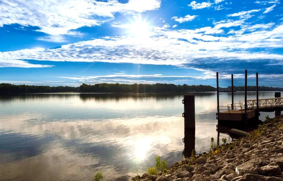 A beautiful view of the Mississippi River with a dock, rocks along the shore, and sunlight reflecting off the water. The sky above is dotted with clouds, evoking a sense of calm and open space. The setting is ideal for boating, fishing, picnicking, or simply watching the sunrise.