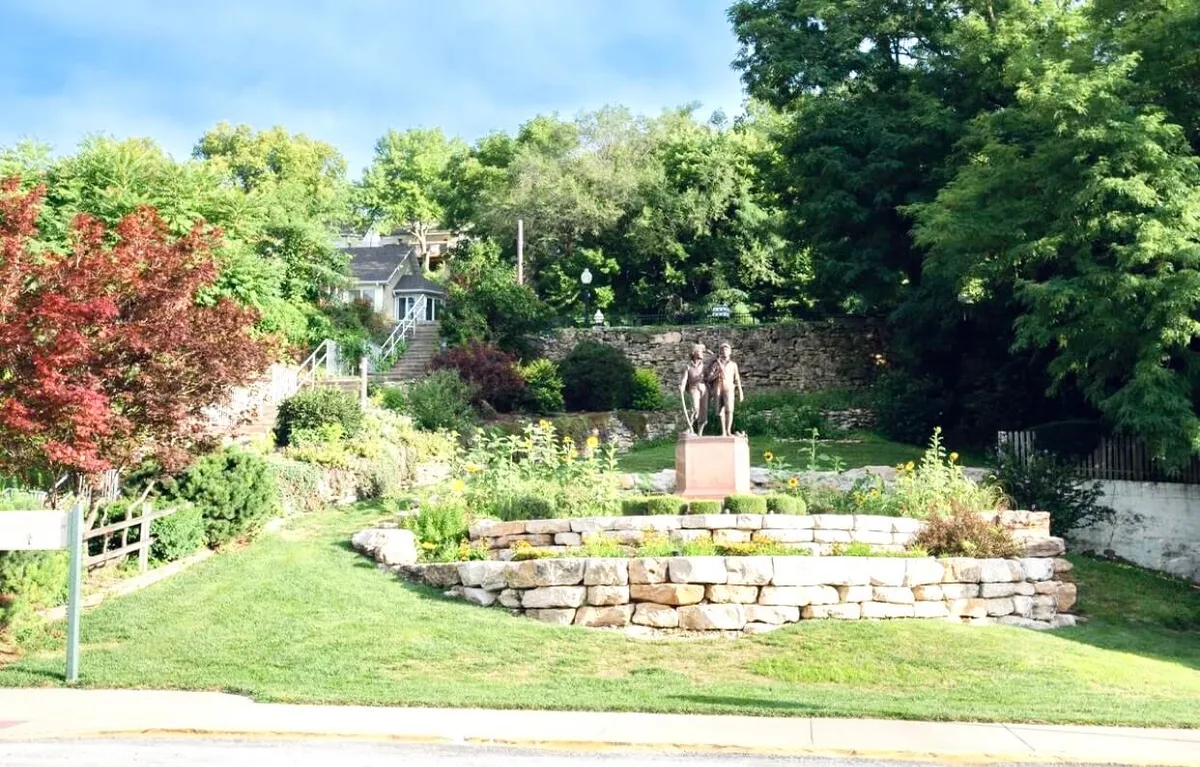 A serene garden scene featuring a sculpture of two figures, vibrant flowers, a stone retaining wall, and lush greenery. The environment encourages strolling, relaxing, and appreciating both local art and the beauty of nature.